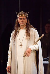 David Tennant with white robes, cross on a chain around his neck, gold scepter in his left hand, gold crown on his head, with his hair cascading down to his breast.