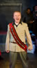 King John in tan uniform jacket and olive pants and a red and black sash across his chest stands front and center yelling; behin him to his right, the Cardinal is in red cardinal robes with black stole; to John's left King Philip in blue jeans, blue shirt and blue tee. The platform with bordered by chain link fences is at the back of the stage, sandbags at the base, and other cast members lined up against the backstage wall.