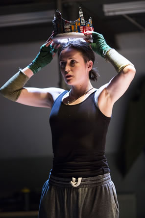 Hal in black tanktop and gray sweats, with green fingerless gloves and an olive green sleeve on the elbow of her right arm, lifts a crown made of aluminum drink cans onto her head.