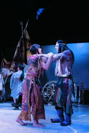 Troilus with long black hair and a headband, wearing  striped, open shirt, multicolor pants to his shins and animal skin boots has his arms on Cressida's shoulders as she grasps his arms: she is in multi-color, doublslitted buckskin dress with tassles, pigtails and bare feat. In the background is a teepee, wagon wheel, and Pandorus in a polka dot blue shirt and long pony tail, his back to us.