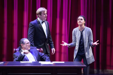 Scalia in a black tux sits at his desk with Brad, also in a tux, standing behind them, both looking past Cat in a long, black-and-gray-striped sweater and dark gray blouse and pants, her hands outstretched in agitation. 
