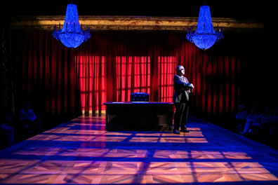 Scalia in a dark suit and tie stands at one end of his large oak desk with leather chair  at the center underneath one of two chandeliers on a stage with the light coming through paned windows casting shadows on the parquet stage and red curtain in the background.