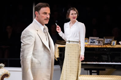 Teck in cream jacket and tie with white shirt stands attentively as Marthe in the background smokes a cigarette next to the piano