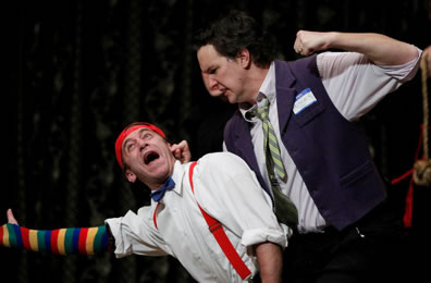 John Harrell as Antiphous, fist raised and holding the ear of Tyler Moss as Dromio: Harrell in vest and tie with a "Hi I Am Prisoner" convention badge, Dromio in red suspenders, bowtie, and multi-color stocking on his right arm