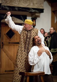 Shylock in long Venitian gown and yellow cap with upraised knife behind Antonio baring his chest