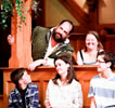 Benedick in green renaissance vest jacket over white shirt leans out from behind a vine-covered post next to two laughing women with three kids on the gallant stools in front of him, one boy looking up at him.