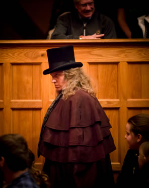 Scrooge next to a wood wall with audience around him, he's wearing a brown multi-layered topcoat and a black tophat