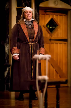 Scrooge in white nightcap, a browsn felt robe with satin lapels and cuffs, tied with string at the middle, over his three piece suit, looking at a crutch leaning against a table in the foreground.