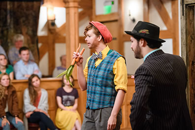 Production photo by Marek K Photography of Dromio of Syracuse eating two carrots as Antipholus of Syracuse watches.