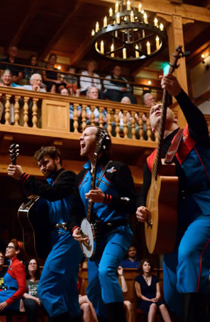 The three wear blue jumpsuits with red trim, and the audience of the wooden Blackfriars is sittein on the stage and in the galleries in the background.