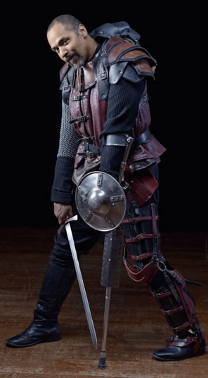 Richard dressed in red and black leather over chain mail armor, a single cructh with a small shield on his left hand, his sword in his right hand pointed down to the floor, his head tilted forward from the armored hump in his back