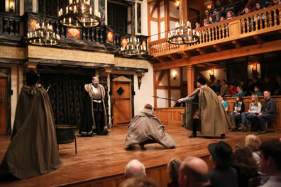 The Ghost in long robe, armored plate, and crown stands at the center of the stage as the soldiers on either side point their spears and Horatio crouches in the middle. The fire kettle is to one side, and the full stage area is in view, with audience in front and to the side and on stage, and chandeliers hanging overhead.