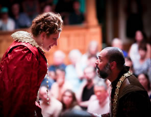 Lear on his knees in brown shirt and gold chain around his neck but bareheaded as Regan in red Jacobean dress, string of pearls, and lace collar bends over looking at him; audience in background.