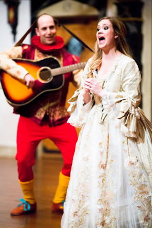 In the background, Accius plays guitar, the strap going over his head, in red pants, brocaded jacket, yellow stocking, and ruf collar, in the foreground, Silena sings, in a gorgeous gold-embroidered white gown