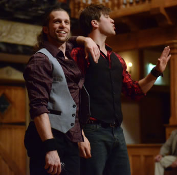 Mercutio in light purple vest and purple shirt with his hand on Romeo's shoulder, in black vest, red plaid shirt, hand up as he exclaims to the audience