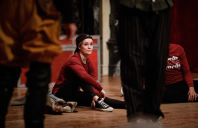 Photo of players sitting on the stage wearing red t-shirt, black pants, and high-tops plus a backward hat, framed by the legs of Rosencrantz and Guildenstern