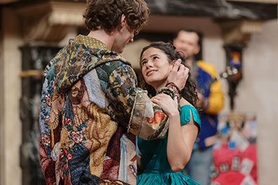 ASC-Romeo_and_Juliet_2025-12 Romeo with back to us is wearing a multicolor hoodie, his right hand stroking her hair as she gaes up into his eyes, her left hand on his right arm. She's wearing a turquoise dress. A man in a blue and yellow letter jacket is playing guitar at the back of the stage.