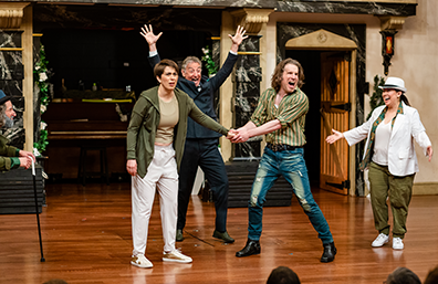 Petruchio in striped shirt and torn pants holds Katherina's left hand with both of his. He's shouting, she, in hooded sweater and sweat pants, is looking dumbfounded. Behind them, Baptista in a suit has his hands raised, to the right Tranio in panama hat and jacket has her arms outstretched, and to the left Gremeo is leaning on his cane.