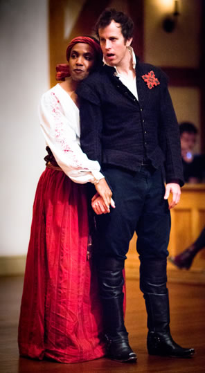 Flamineo stands in blue rennaissance short jacket, trousers, and kneehigh black boots with a red badge on his left lapel, and Zanthe in simple white blouse with floral embroidery and floor-length pleated red skir rests her head on the back of Flamineo's shoulder and holds his hand. 