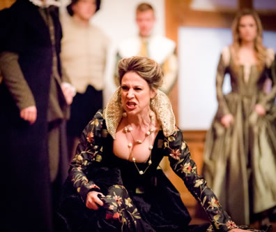 Livia in black dress with floral sleeves and skirt and gold lace high collar screams as she kneels on the floor, other characters standing in the background
