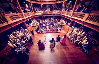 Four musicians in custom in the center of the wood stage in a wood, oval shaped theater of two levels, filled with people.