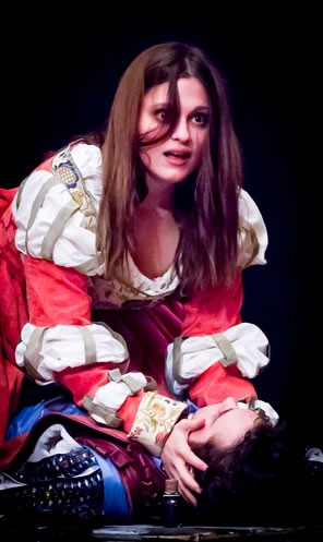 Juliet in red and white Elizabethan dress, hair hanging over her face looking hotter than hell sits on the dead Romeo with her hands clasping his head.