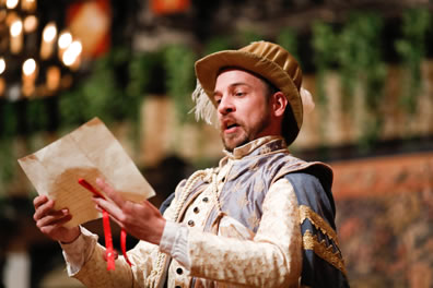 Boyet is wearing a satiny cream-colored jacket, blue and gold cape, and gold courtier hat with a feather sticking out as he reads the letter (with red ribbon seal) he holds out in front of him with both hands.