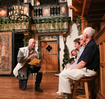 Ferdinand, dressed in white cloak over silver courtier knee breeches, kneels on one knee, paper in hand, looking at men and women sitting on stools next to a wood wall on the edge of the stage. In the background is the Blackfriars black-trimmed tiring stage, with wood door, a tapestry, and vines handing off the balcony trim.
