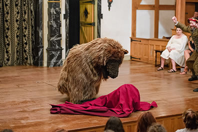 Production photo by Marek K. Photography of the bear sniffing at the blanketed baby with Antigonus on the right side waving his arms.