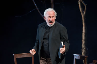 Simon Callow, book clutched in hand, with two chairs and tree behind him