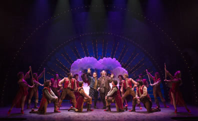 Production number, with neon-lighted and feathered arch in the background and the Elizabethan-costumed dancers around Nick Bottom and Nostradamus, waving in the center.