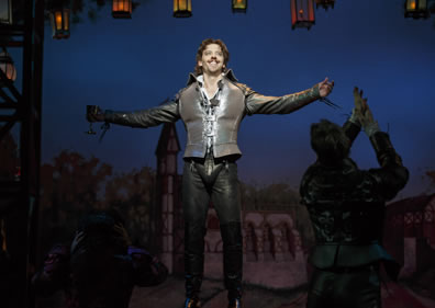 In gun metal gray leather jerkin, shirt, pants, and large codpiece, Shakespeare stands with arms outstretched--goblet in right hand--taking in the adulation of the crowd (we can see a silhouette of an applauding fan to the right)