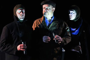 Actors in overcoats and stocking hoods hold flashlights casting lights onto their faces
