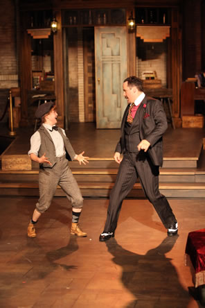 Moth in newsboy cap, brown vest and brown knee-pants with stockings and bowtie dancing with Don Armato in dark gray pinstriped three-piece suit with red-dotted cravat and rose boutineer; the speakeasy set is in the background.