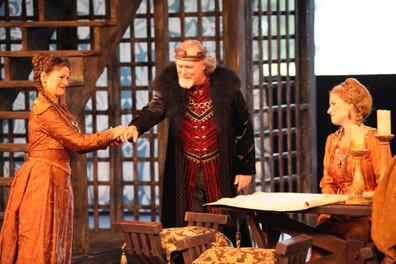 Lear, in red royal doublet, chain around his neck, bronze crown on his head, and a fur overcoat holds hands with Regan who, like Goneril, is wearing gold, formal gowns, their hair done up in braids. Seats with embroidered cushions, candelabra on the table, and a latice-work background make up the set