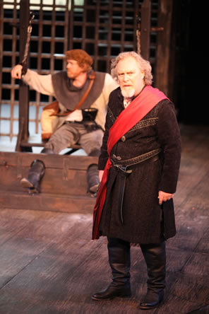Lear in knee-length coat, red sash across his left shoulder and tied at a knot at his right waist, black riding boots, Kent in linen work shirt, rough pants, a knit work collar and brown peasant cap, his booted calves int he stocks, his arms hung by the wrists in irons on chains