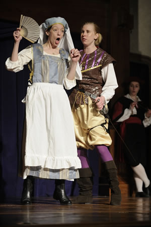 Nurse in blue dress and head dress and white apron fans herself and her mouth is agape as Mercutio in jeweled vest, gold pants, purple stockings and boots and hand on sword hilt stands right up next to Nurse. Peter is in black with red sash and white stockings crossed leg leaning against a wall in the background.