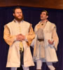 Leontes to the right with hands cupped and outstretched in exasperation talking to the side of Camillo, who patiently holds his hands folded at his waist, both where white sleeveless tunics with satiny shirts underneath and white leggings, Leontes in a crown.