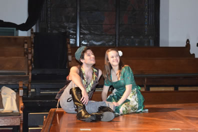 Florizel and Perdita sit on a stage floor, with church pews in the background. he's dressed in flower brown vest, gray work pants and brown boots and wears a simple gray cap. She's wearing a floral green dress and has a white carnation in her hair.
