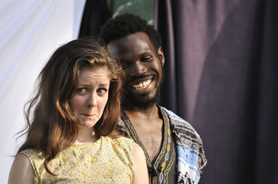 Phoebe with windswept brunette hair and wearing a yellow dress has a frown in her expression while behind her, Sylvius with Bohemian shirt and serape vest smiles broadly at her.