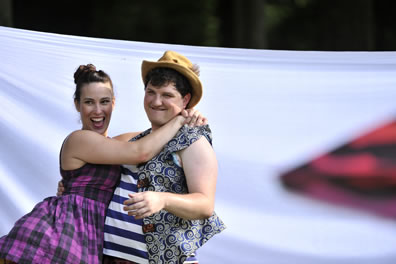 Phoebe in shoulderless purple plaid dress that billows out at the waist has her hands clasped around the neck of Touchstone, who is wearing a blue and white striped t-shirt, blue-patterned vest, and straw fedora.
