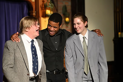 Production photo of Hamlet with his arms around Rosencrantz and Guildenstern, both in gray suits.