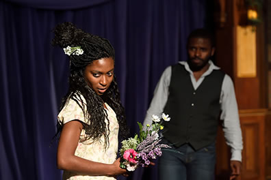 Production photo of Ophelia with flowers, looking away with a sly expression. Laertes is in the background.