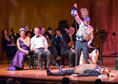 Hippolyta in purple gown and crown sits next to Theseus in white suit shirt and bowtie in front of members of the orchestra string section, all in black formal wear, leaning over to see, bottom in whicker breastplate and funny helmet lies on the floor as Moonshine in flack-jacket, baseball cap with thorn bush, holding up a lantern and leash on white dog stands over him.