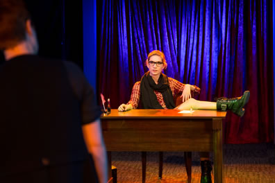 Mephistopholis, wearing a brown checked shirt dress, boots, glasses, and a sweater scarf around her neck, sits at Faustus's desk,  one leg up on the desk top, as she talks with Faustus, whose back is to the camera.
