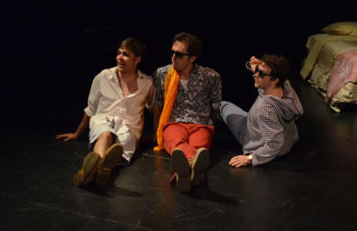 Sitting on the floor are Romeo in white shirt, unbuttoned to the chest and white shorts, Mercutio in sunglasses, orange pants, purple print shirt and orange scarf, and Benvolio, also weareing sunglasses in a blue striped hoodie and shin-length pants