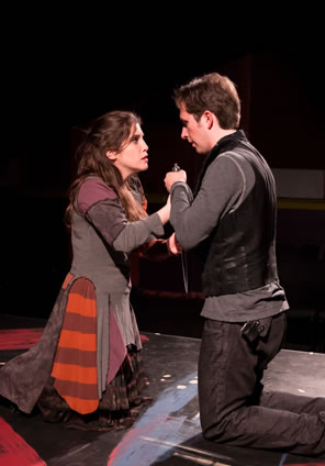 Annabella in gray tunik, purpe sleeves and red-striped panels of fabrick over a black print dress kneels opposit Giovanni in black leather vest, black jeans, and grey sweatshirt, both clasping a dagger between them.