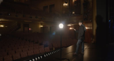 Mike, in hat, coat, scarf, and jeans is standing on a stage, left hand in jacke pocket, right hand stretched out as an actor speaking, with a work lamp in the middle of the picture and the theater seats in the background and to the left