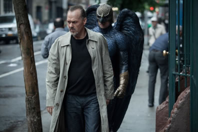 Keaten in black sweatshirt, blue jeans, gray trenchcoat walking down the street (tree on his right, gate on his left) with Birdman in black armor, gold gloves and hood/mask, and large black wings behind him