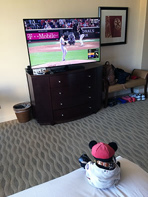 Teddy bear in Nationals uniform sits on a hotel bed watching the baseball game on a TV perched on a dresser across the room.